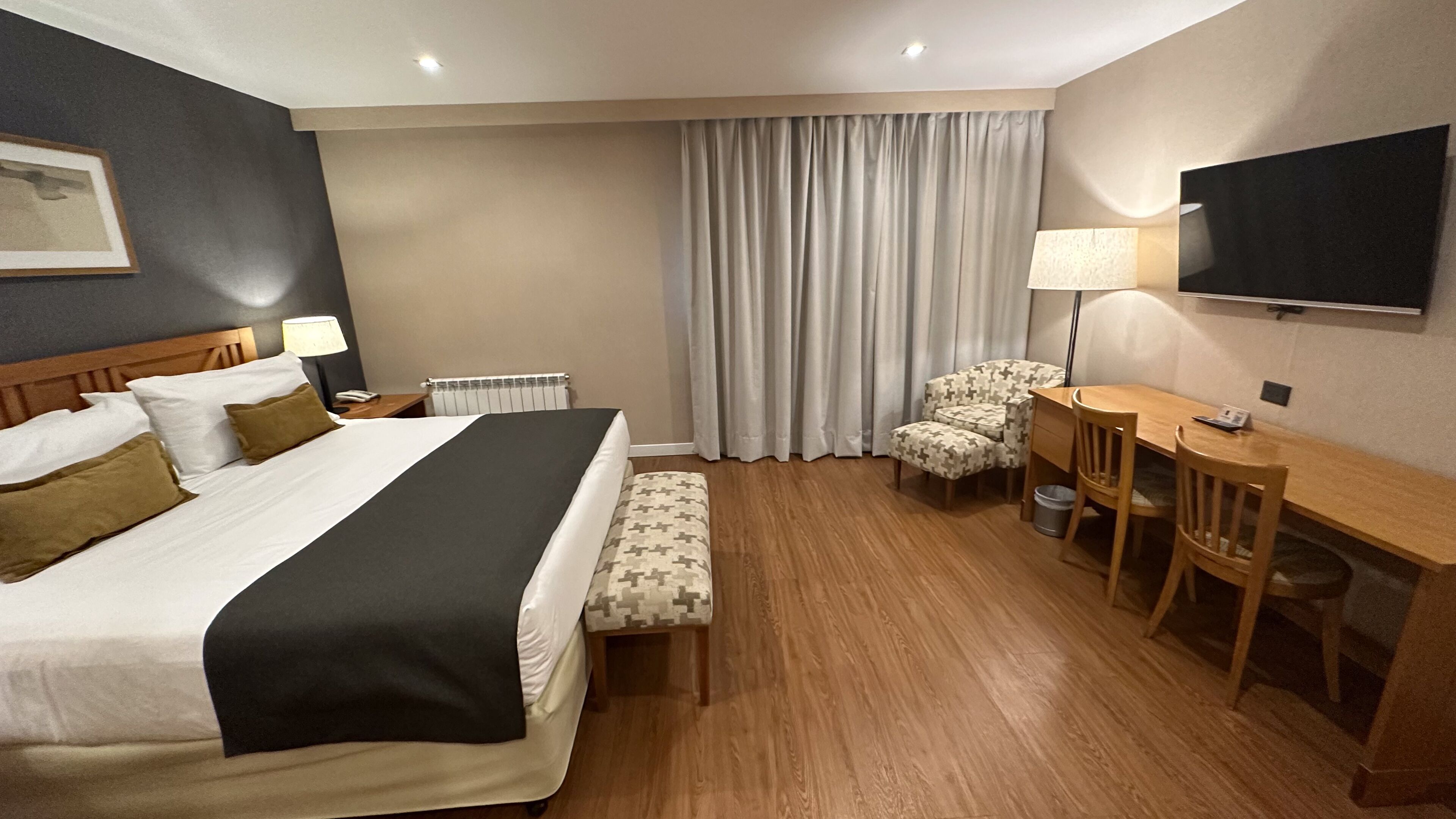 A hotel room with a large bed, a desk with a chair, a television, a window with curtains, and a radiator.