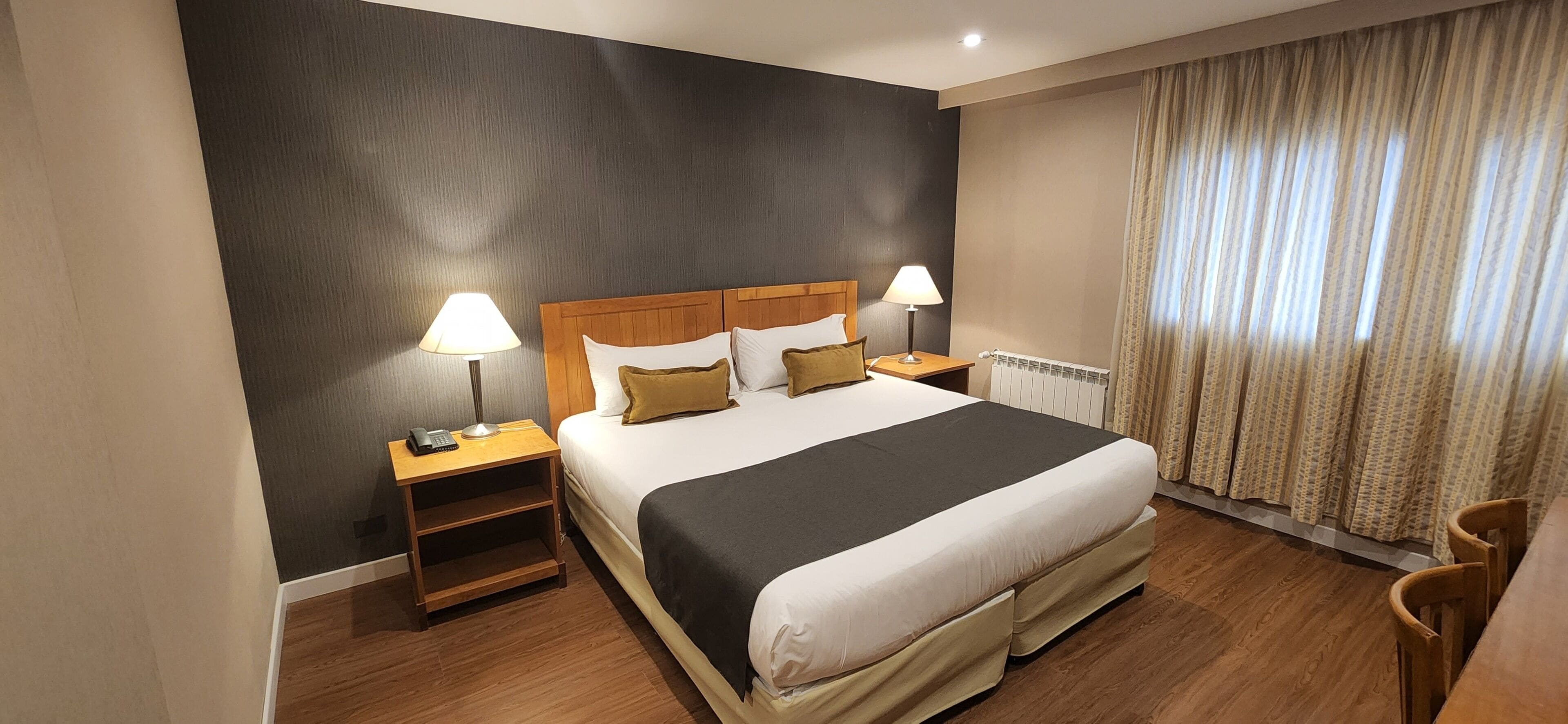 Une chambre d’hôtel avec un grand lit, deux tables de chevet avec des lampes, une tête de lit en bois et une fenêtre avec des rideaux.