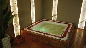 Tinaja rectangular con base de madera y grifo metálico, ubicada en una habitación con piso de madera y un ventanal grande.