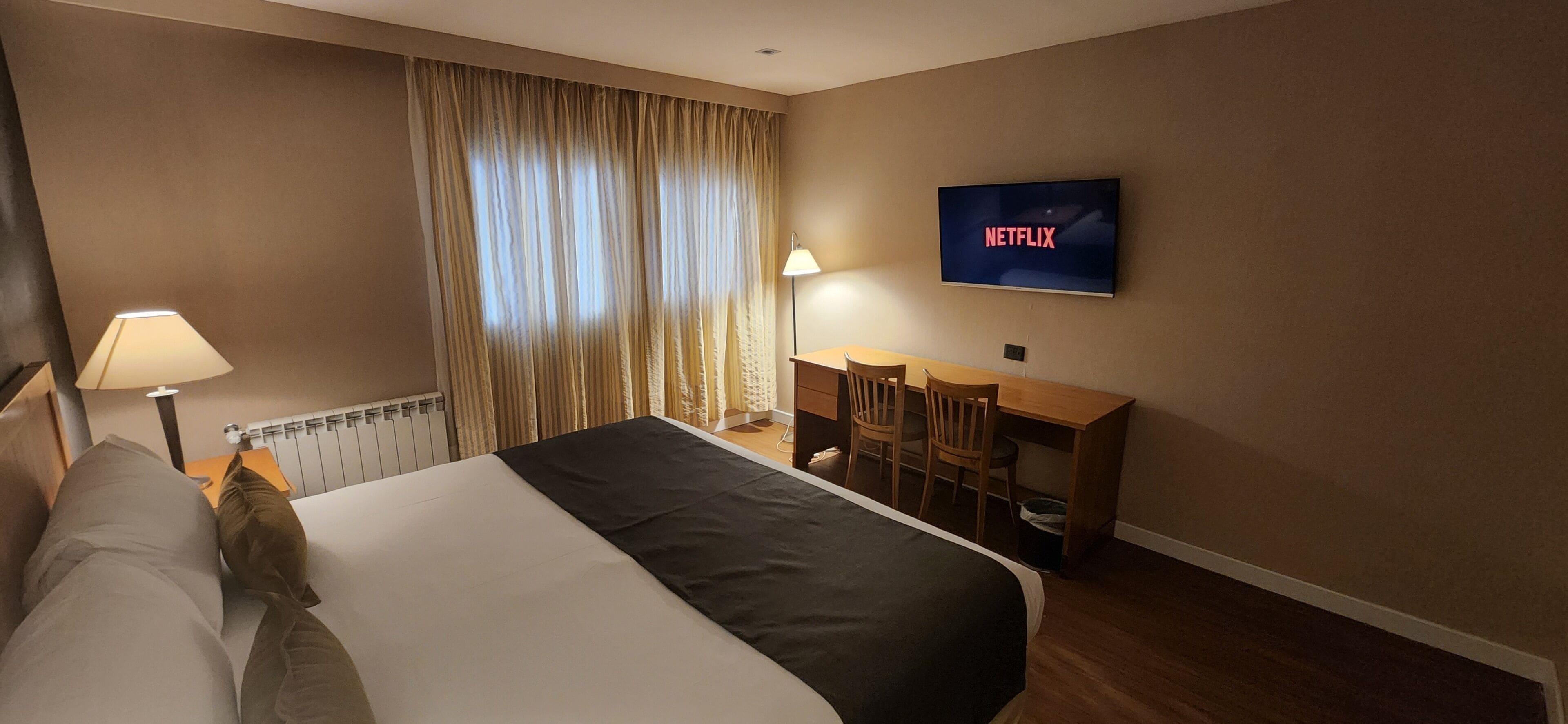 Habitación de hotel con una cama, un escritorio, una silla, un televisor mostrando 'NETFLIX' y una lámpara.