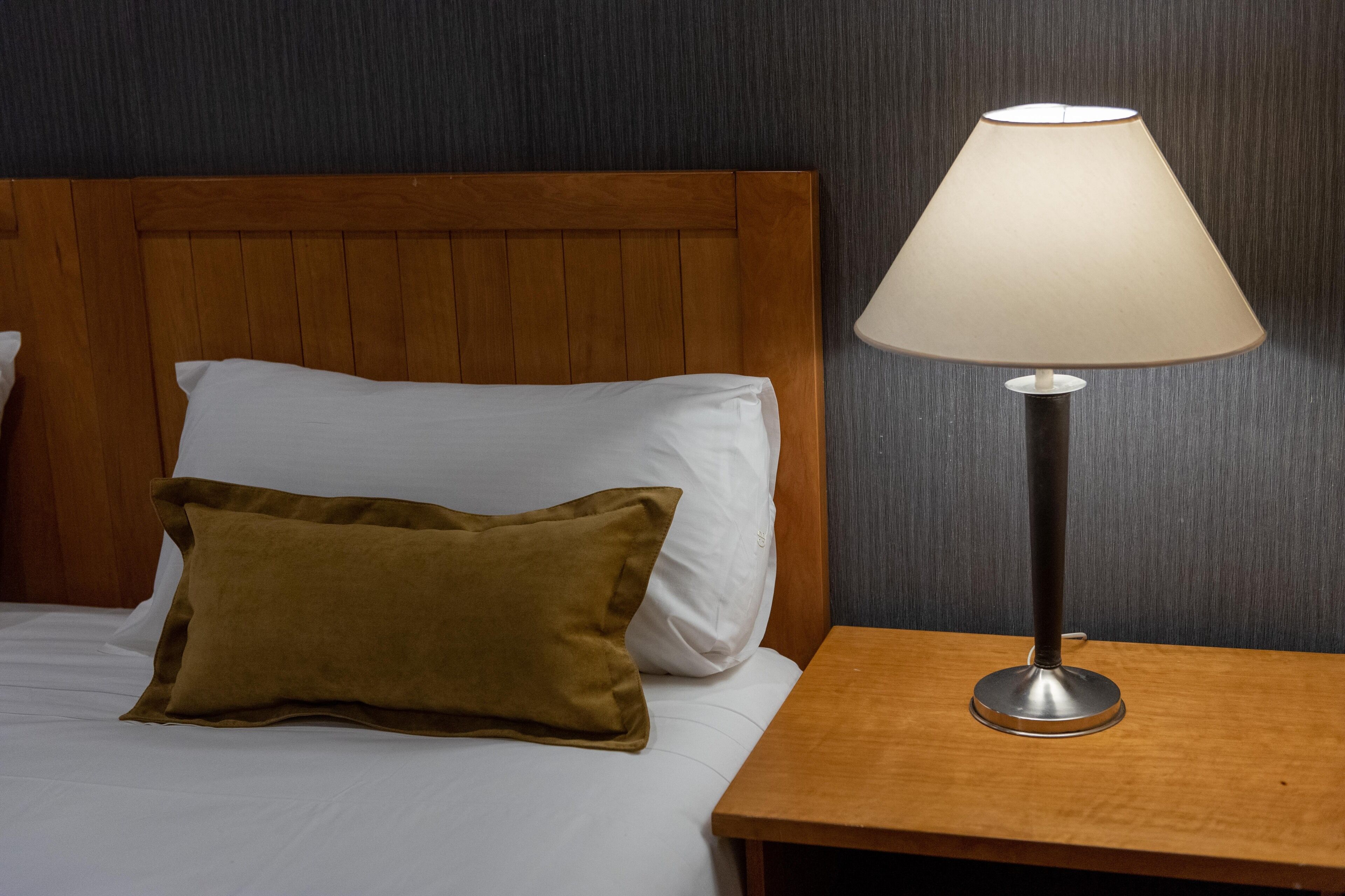 Un lit bien fait, avec une tête de lit en bois, un oreiller marron et un oreiller blanc. Une lampe de chevet à abat-jour blanc et à base métallique est posée sur une table de chevet en bois, à côté du lit.