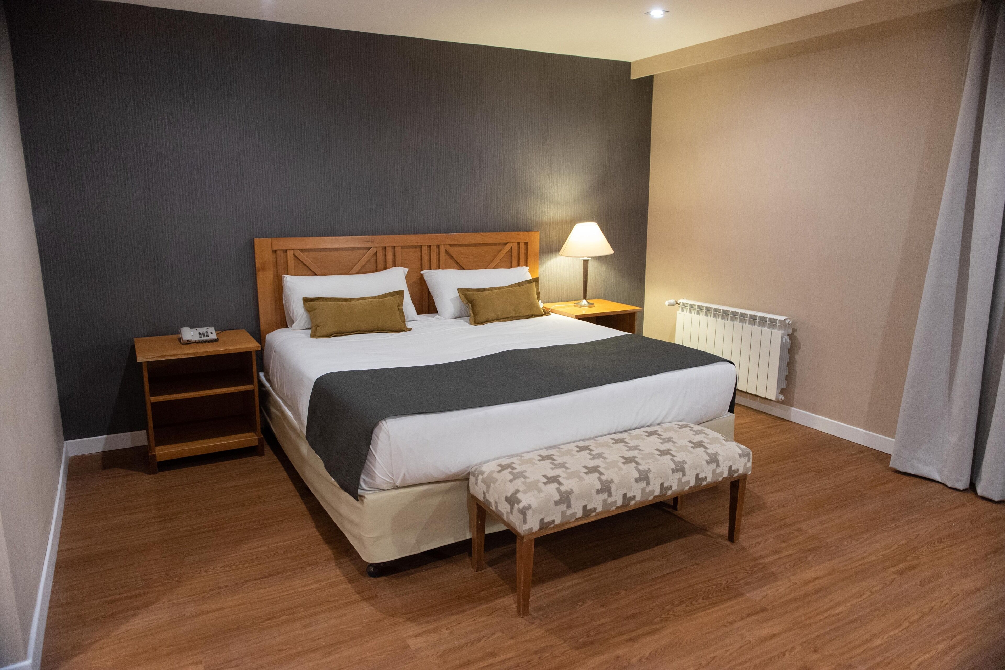 A hotel room with a large bed, a bedside lamp, a wooden nightstand, a bench, and a radiator.