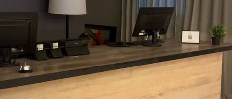 Una mesa de recepción de hotel con mostrador de madera, dos monitores de computadora, una lámpara de escritorio y una pequeña planta en maceta.