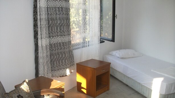 Una stanza piccola e pulita con un letto singolo, un comodino in legno, una sedia e una finestra con tende.