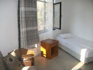 Una habitación pequeña y limpia con una cama individual, una mesita de noche de madera, una silla y una ventana con cortinas.