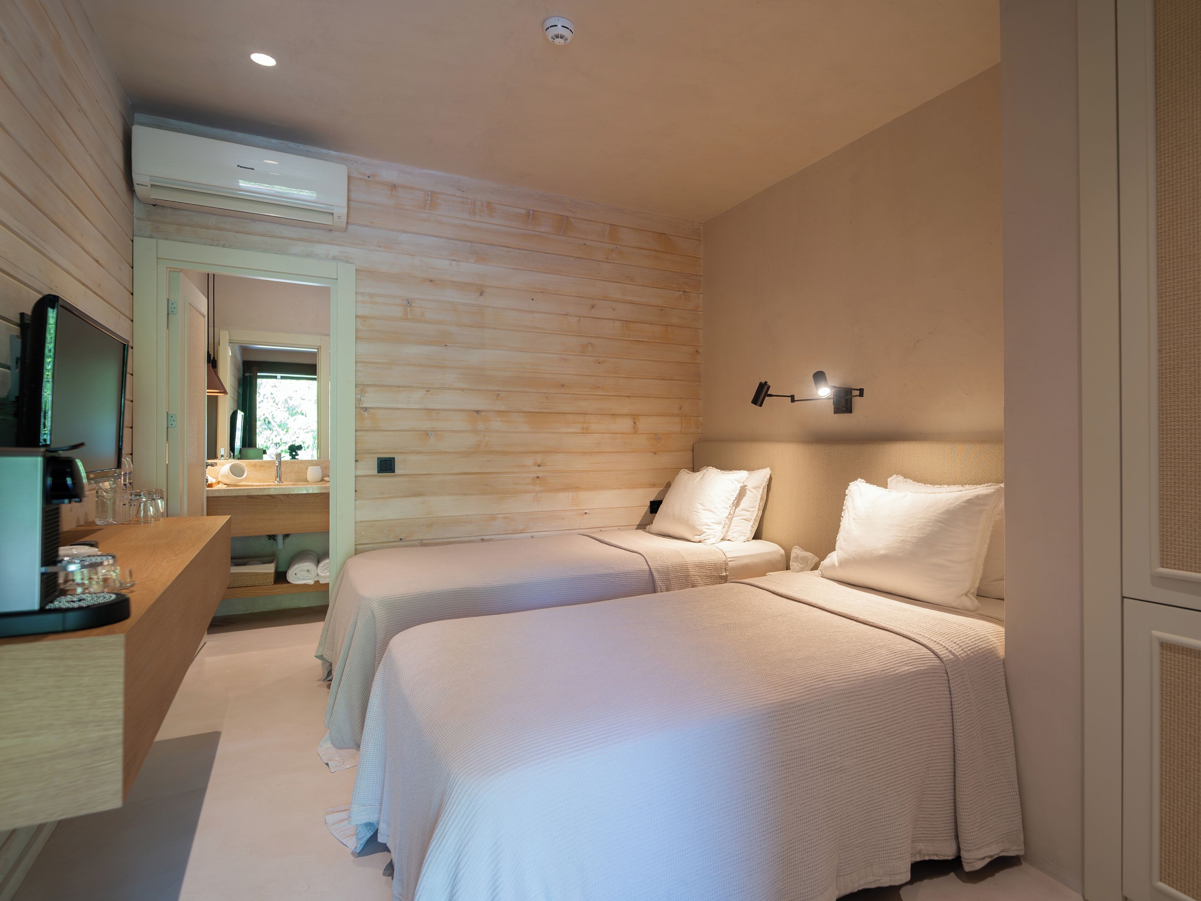 A modern hotel room with two beds, a wooden wall, a small desk with a mirror, and a ceiling-mounted air conditioner.