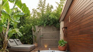 A hot tub with a wooden deck, a seating area with cushions, and lush greenery in the background.