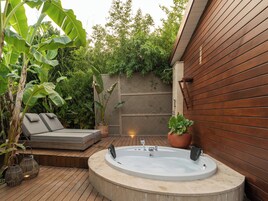 Un jacuzzi con cubierta de madera, zona de estar con cojines y abundante vegetación al fondo.