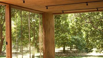 A spacious room with wooden flooring and large windows, offering a view of trees and greenery outside.