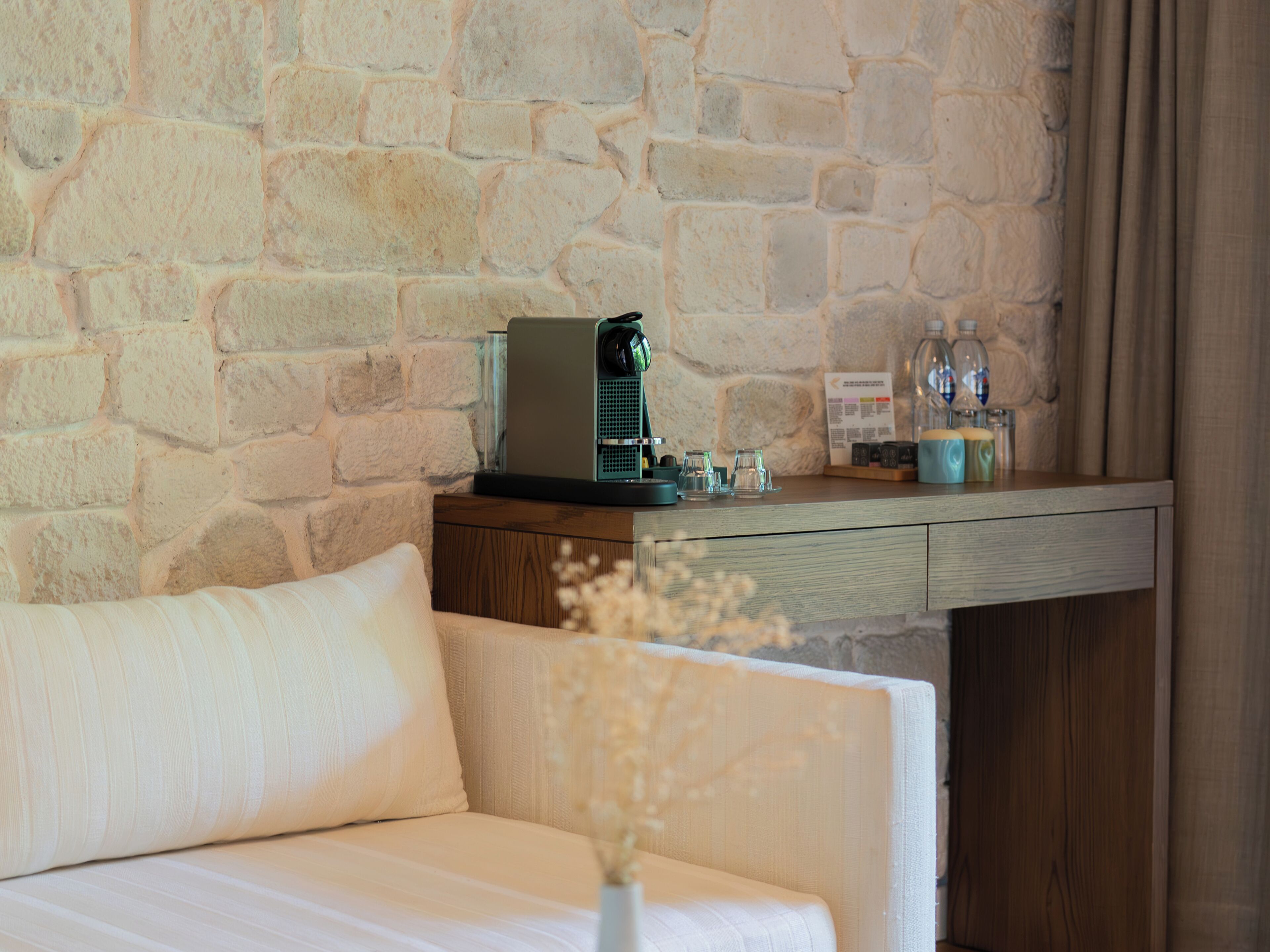 A hotel room with a stone wall, a wooden desk with a coffee machine, a bottle of water, and a vase with dried flowers.