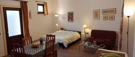 A small, cozy room with a bed, a sofa, a dining table with chairs, and a lamp.