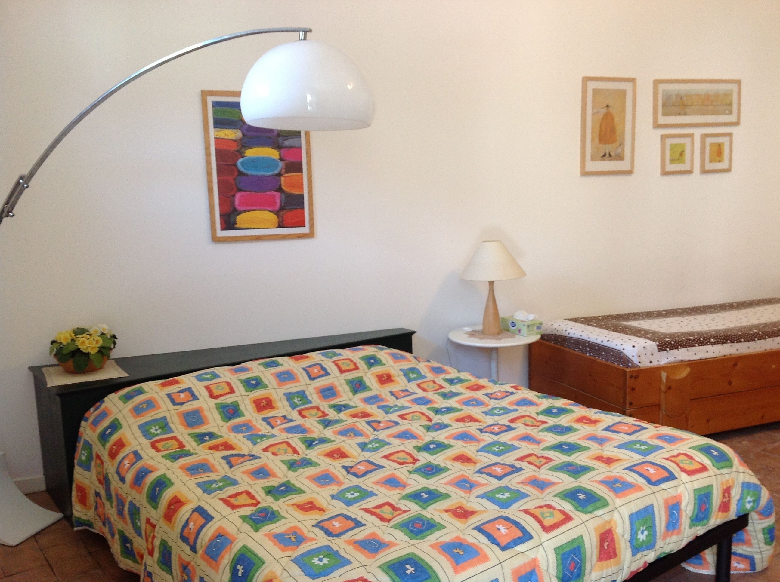 A bedroom with a colorful quilt, a bedside table with a lamp, a small plant, and a framed artwork on the wall.