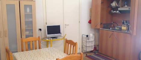 A compact room with a dining table, chairs, a small TV on a stand, a wardrobe, and a kitchen area with a sink and utensils.