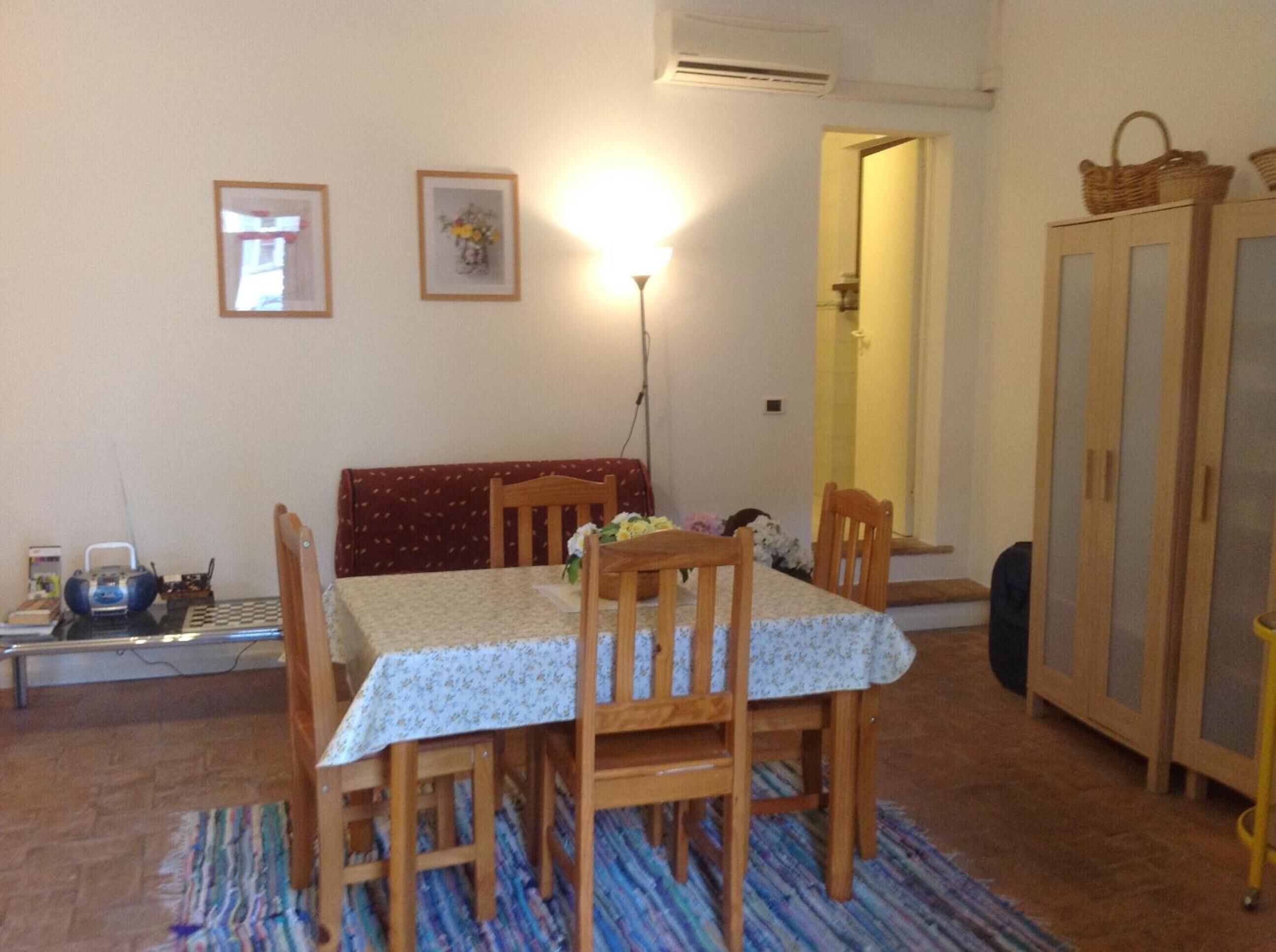 A dining area with a table set for four, surrounded by wooden chairs. There is a bench against the wall, a lamp on the table, and a cabinet with baskets on top.