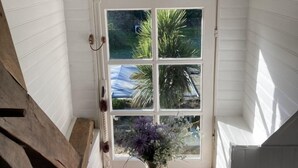 A small, white-paneled room with a sloped ceiling, a wooden bench, a small table with a vase of flowers, and a window with a view of greenery outside. - La Marmoire (Soudan)