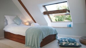 A bedroom with a sloped ceiling, a bed with a wooden headboard, a window with a view of greenery, and a bedside table with a lamp. - La Marmoire (Soudan)