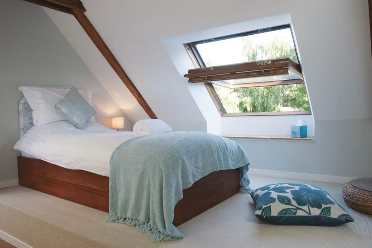A bedroom with a sloped ceiling, a bed with a wooden headboard, a window with a view of greenery, and a bedside table with a lamp.