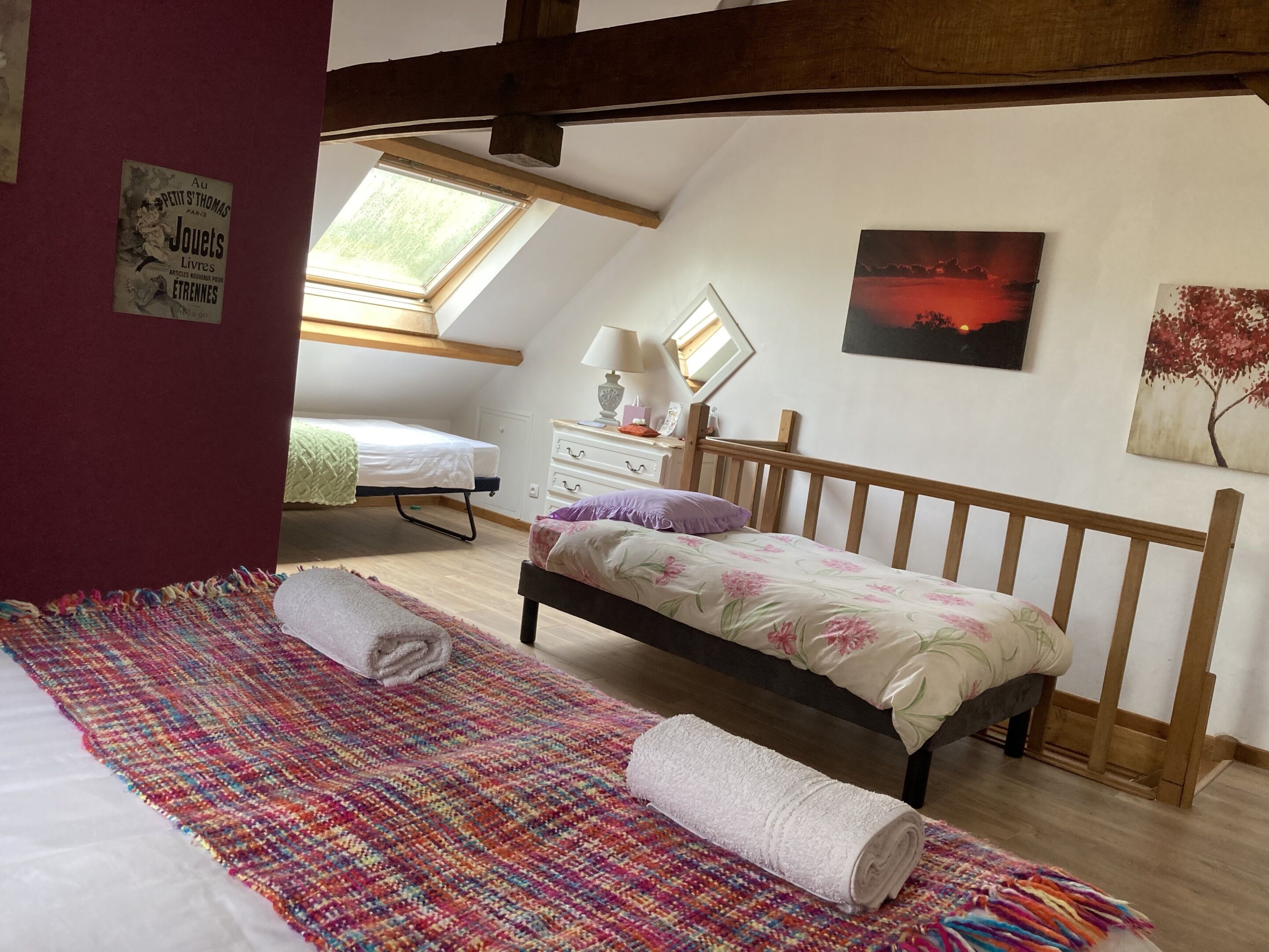 Een slaapkamer met een schuine plafond, een bed met een bloemenbedekking, een kleurrijk geweven vloerkleed, een klein nachtkastje met een lamp en een schilderij van een zonsondergang.