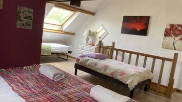 Een slaapkamer met een schuine plafond, een bed met een bloemenbedekking, een kleurrijk geweven vloerkleed, een klein nachtkastje met een lamp en een schilderij van een zonsondergang.