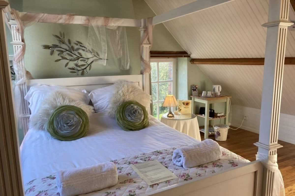 A four-poster bed with rolled towels and a floral quilt, a small table with a lamp, and a window with a view of greenery.