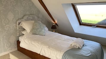 Un dormitorio con techo inclinado, una cama con ropa de cama blanca, una manta azul y una ventana con vistas a la vegetación.