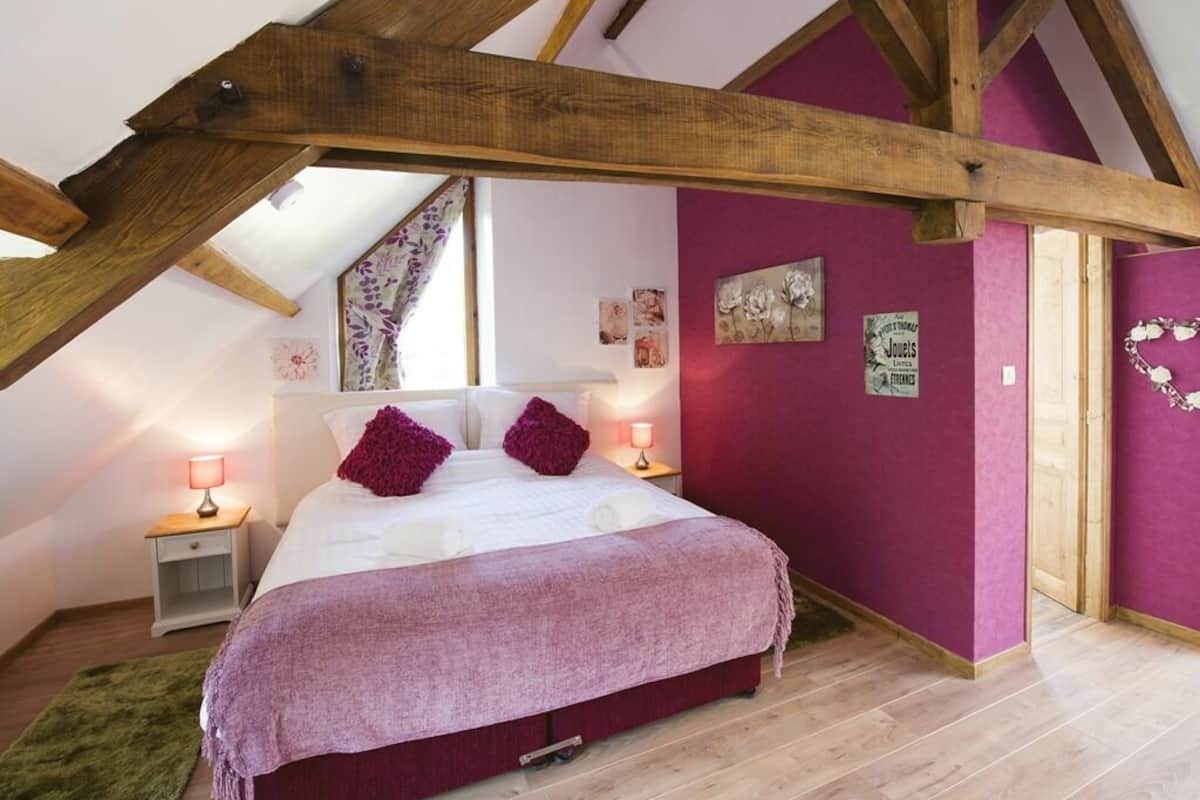 A bedroom with a sloped ceiling, wooden beams, a bed with a purple blanket, two bedside lamps, and a green rug.
