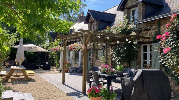 A patio with outdoor furniture, a pergola with hanging flower baskets, and a stone building with a pitched roof. - La Marmoire (Soudan)
