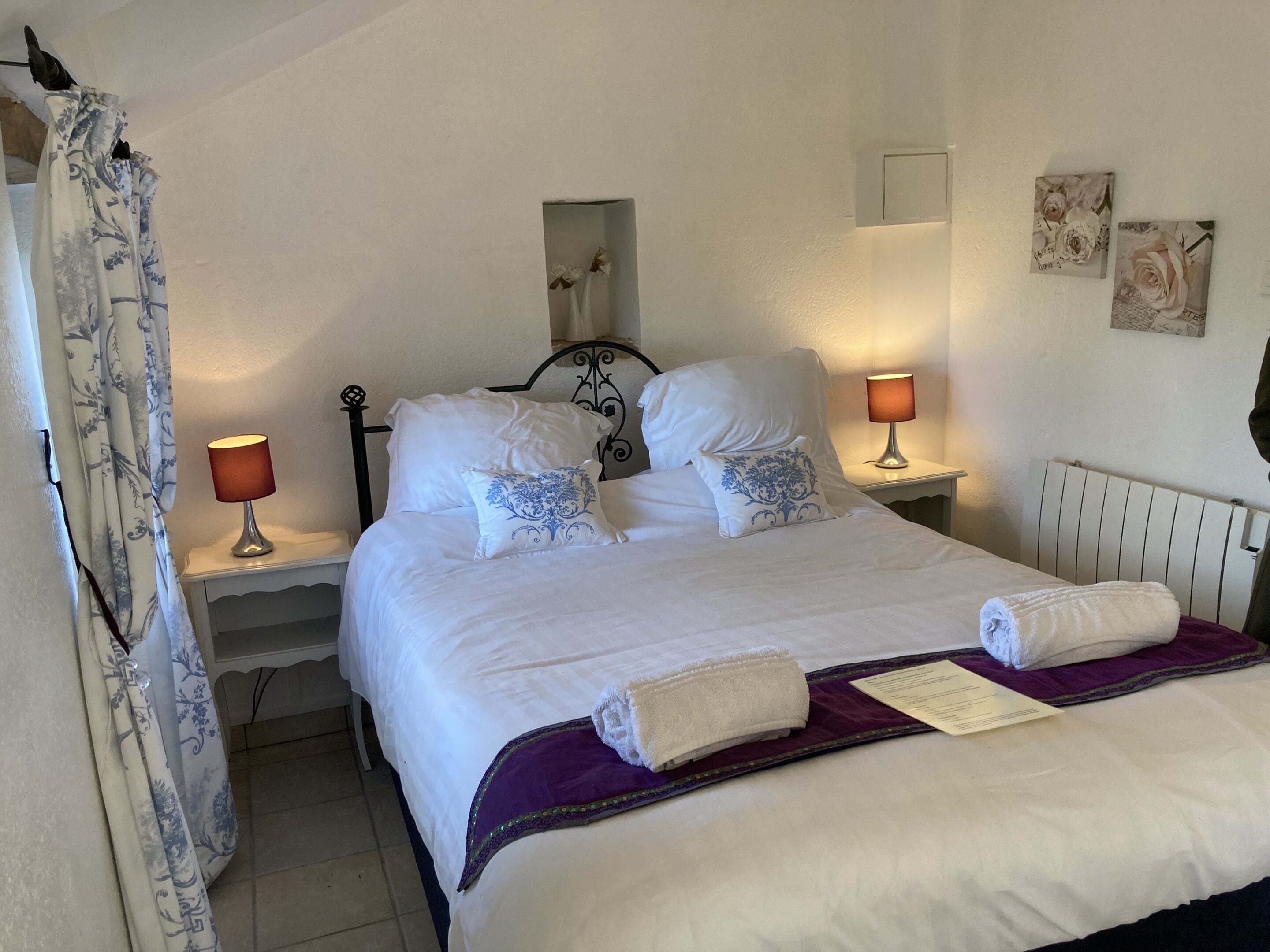 A neatly made bed with white linens and decorative pillows, a purple throw blanket, and a folded towel. There are two bedside lamps, a white nightstand, and a wall-mounted mirror.