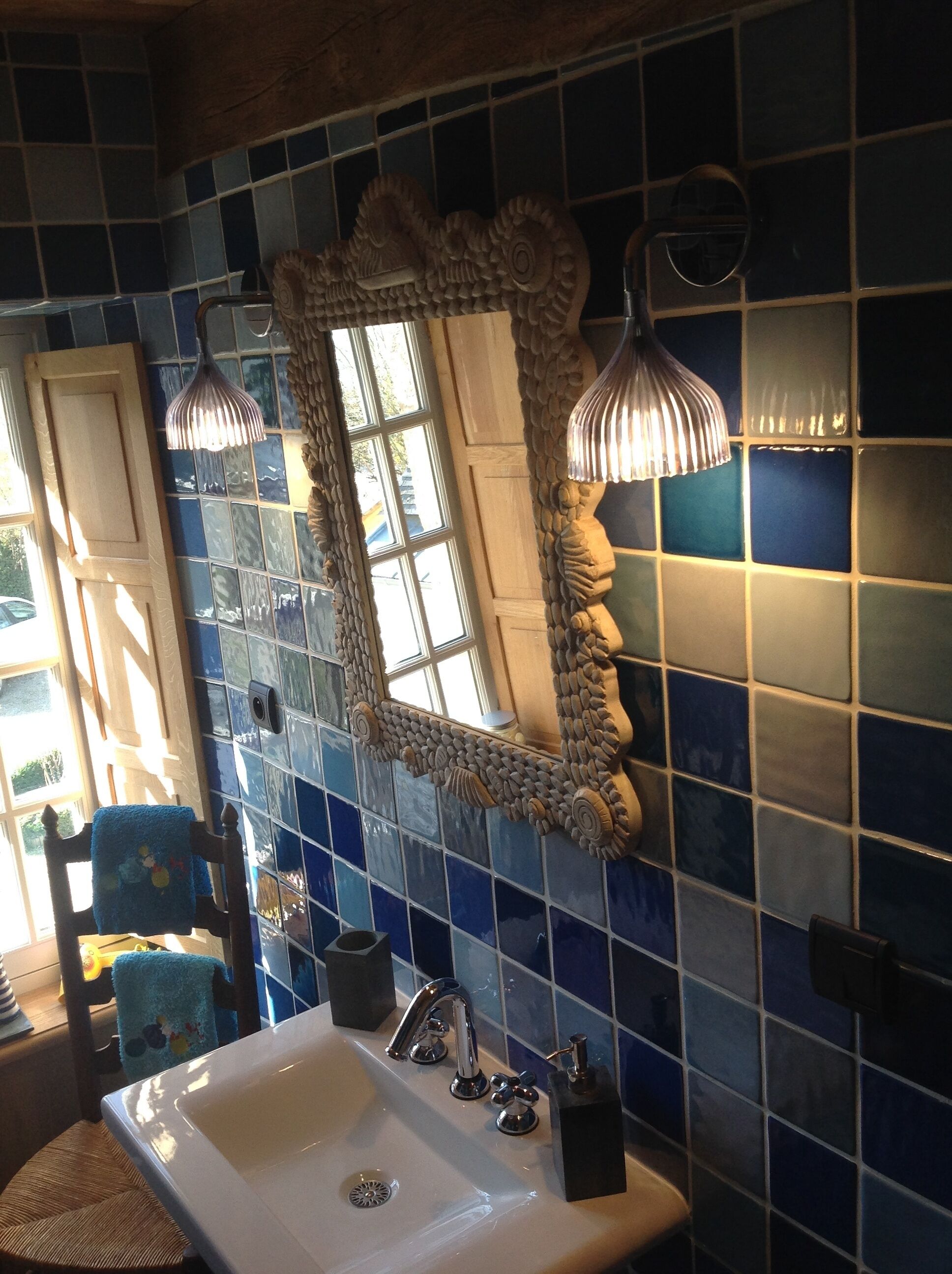 Un baño con un lavabo blanco, paredes con azulejos azules, un espejo decorativo y una ventana.