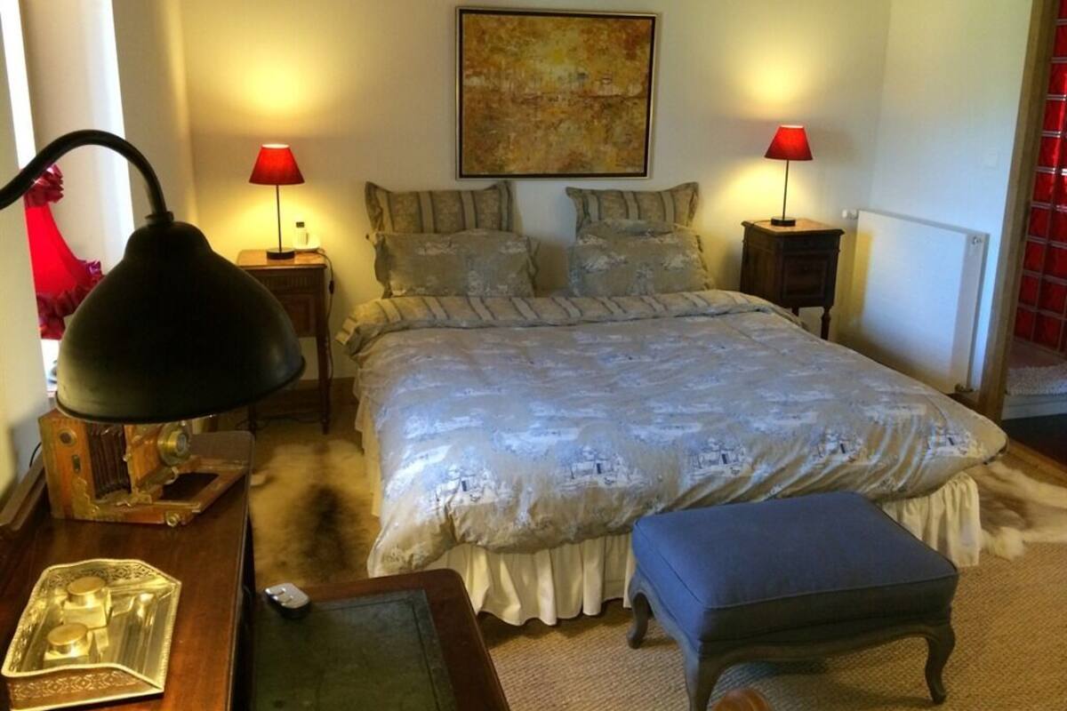Een slaapkamer met een groot bed, twee bedlampjes, een schilderij aan de muur en een klein tafeltje met een lamp en decoratieve voorwerpen.