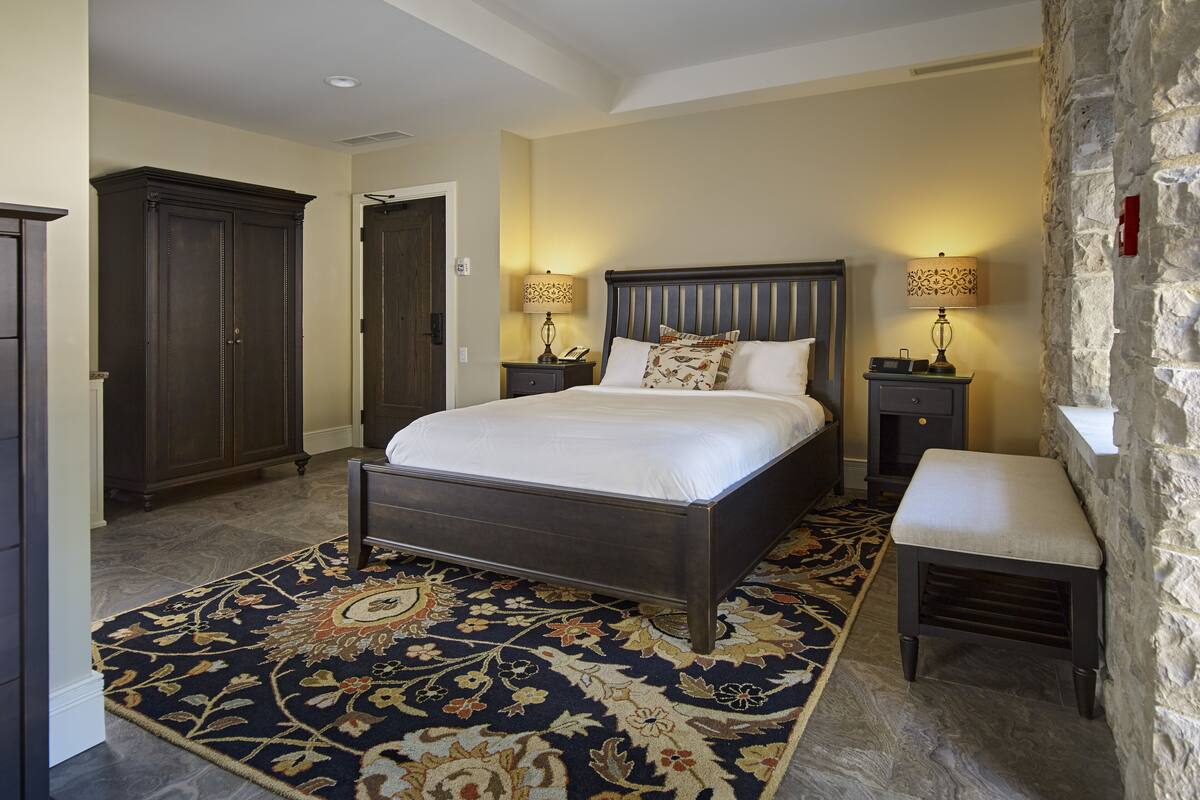 A bedroom with a large bed, two bedside tables, a nightstand, a bench, a carpet, and a wardrobe.