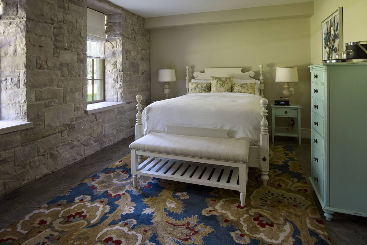 A bedroom with a stone wall, a bed with white bedding, a bench, a dresser, and a patterned carpet.