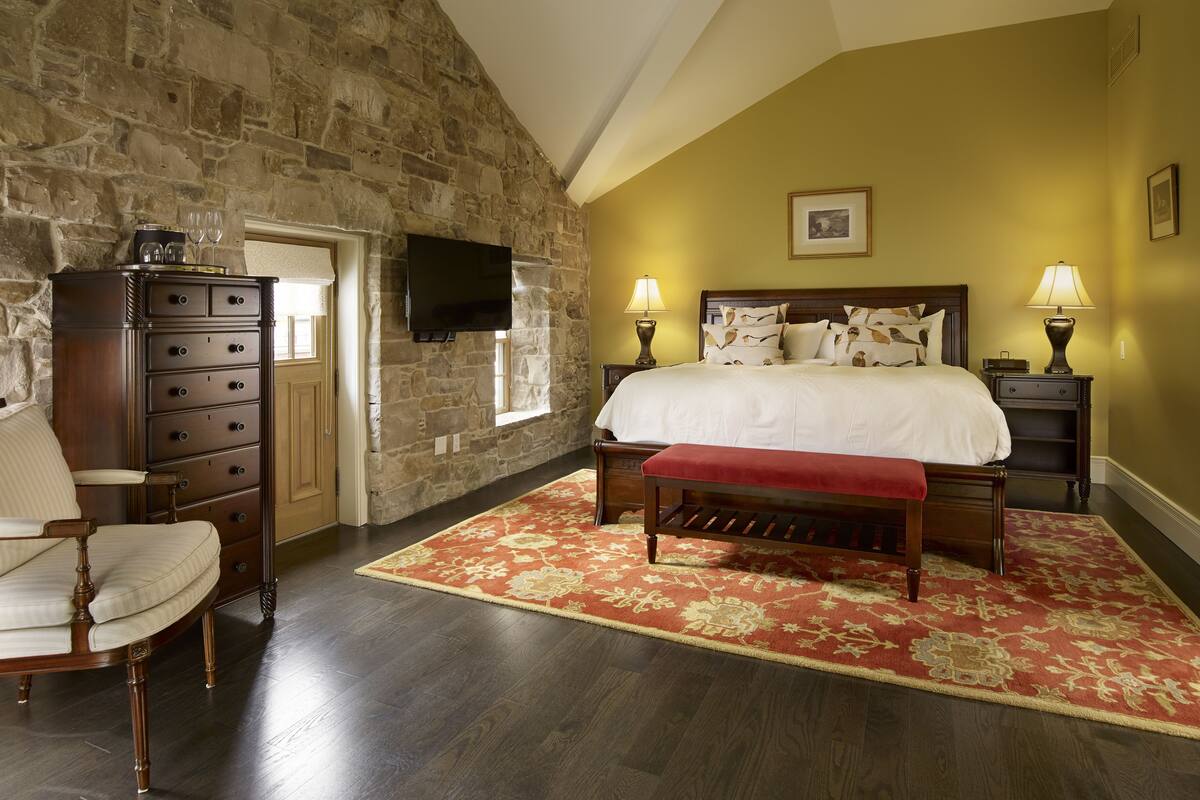 A bedroom with a stone wall, a bed with a red and beige patterned rug, a wooden dresser, a chair, and a flat-screen TV mounted on the wall.