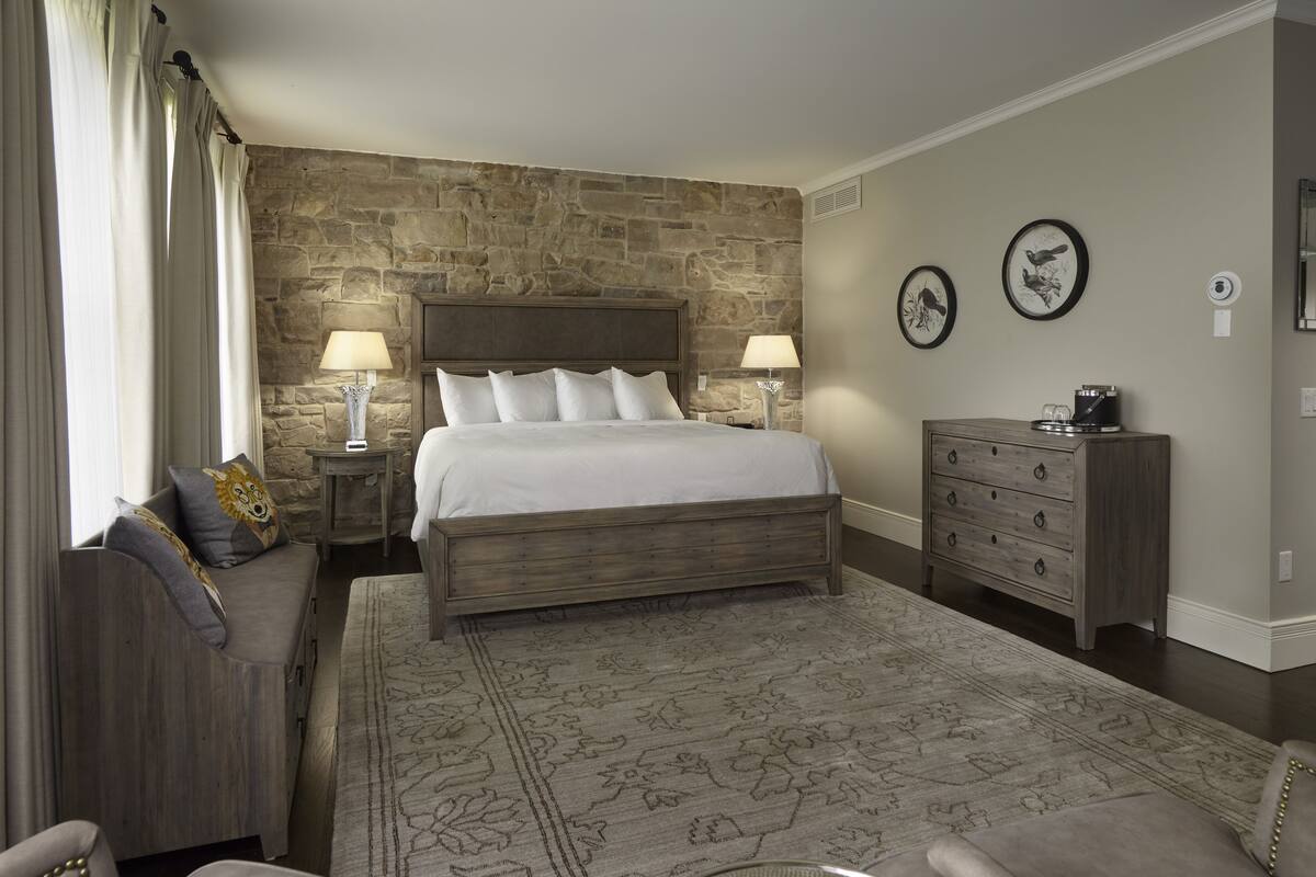 A bedroom with a stone wall, a large bed, two bedside tables with lamps, a dresser, and a patterned rug.