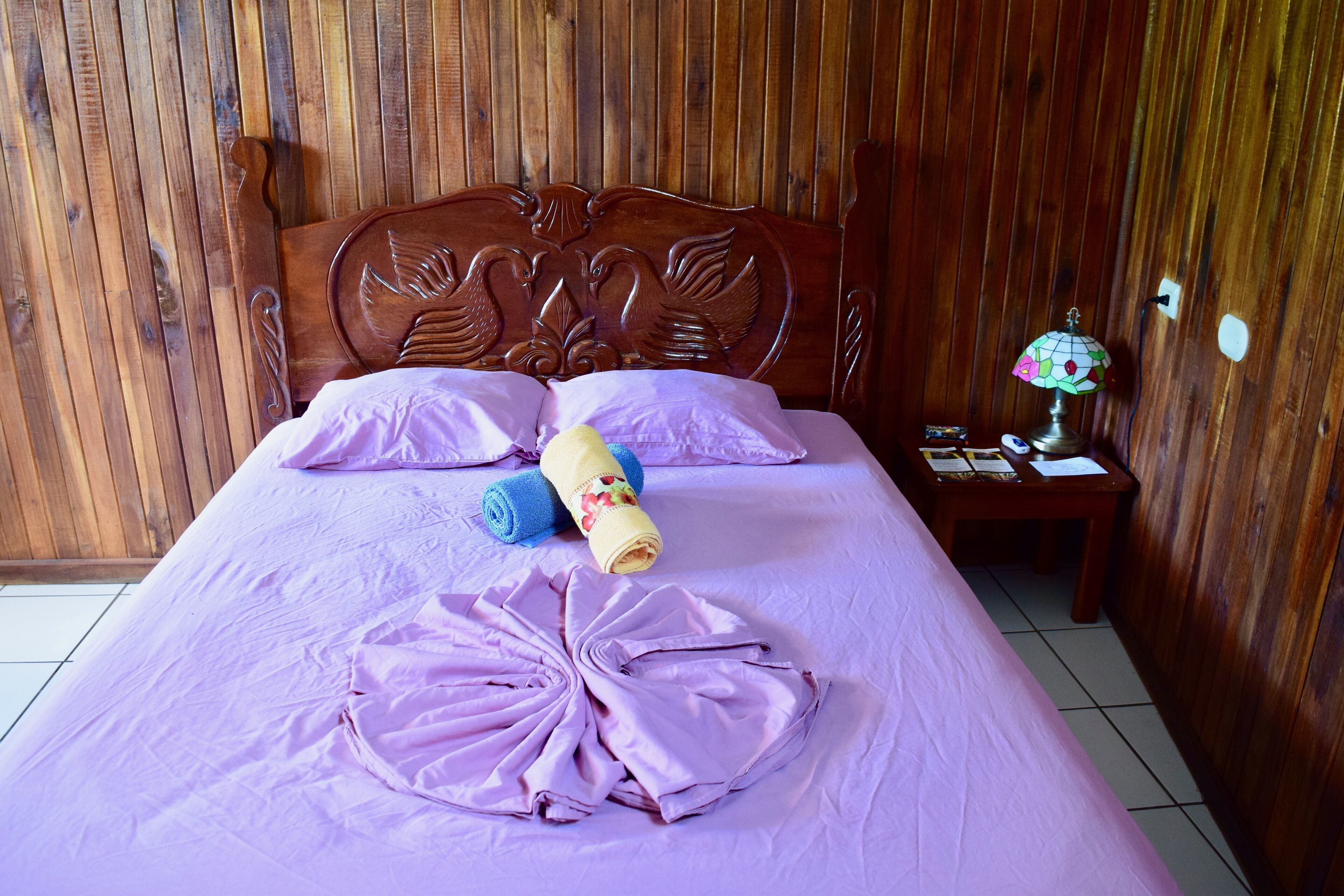 Un lit bien fait, avec du linge de lit rose, une tête de lit en bois et une table de chevet sur laquelle se trouvent une lampe et un livre.