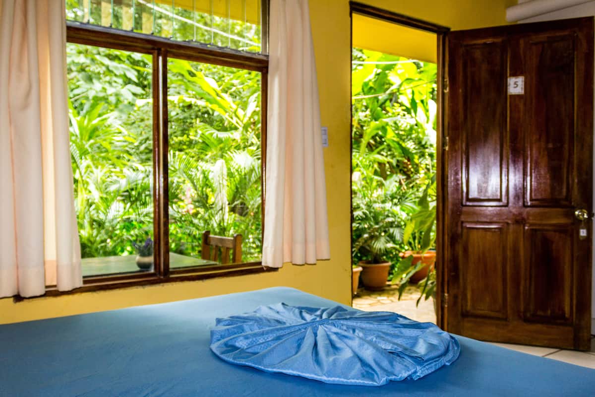 Quarto com cama, janela com cortinas, porta e vista para o verde lá fora.