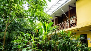Un edificio amarillo con balcón que da a un jardín exuberante con diversas plantas y árboles.