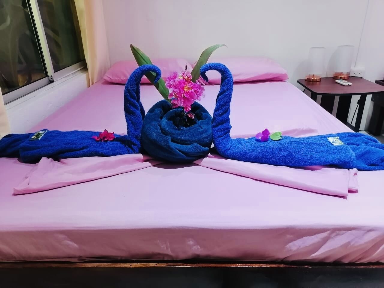 A hotel room with a pink bedspread, a blue towel sculpture, a pink flower, and a small table with a lamp.