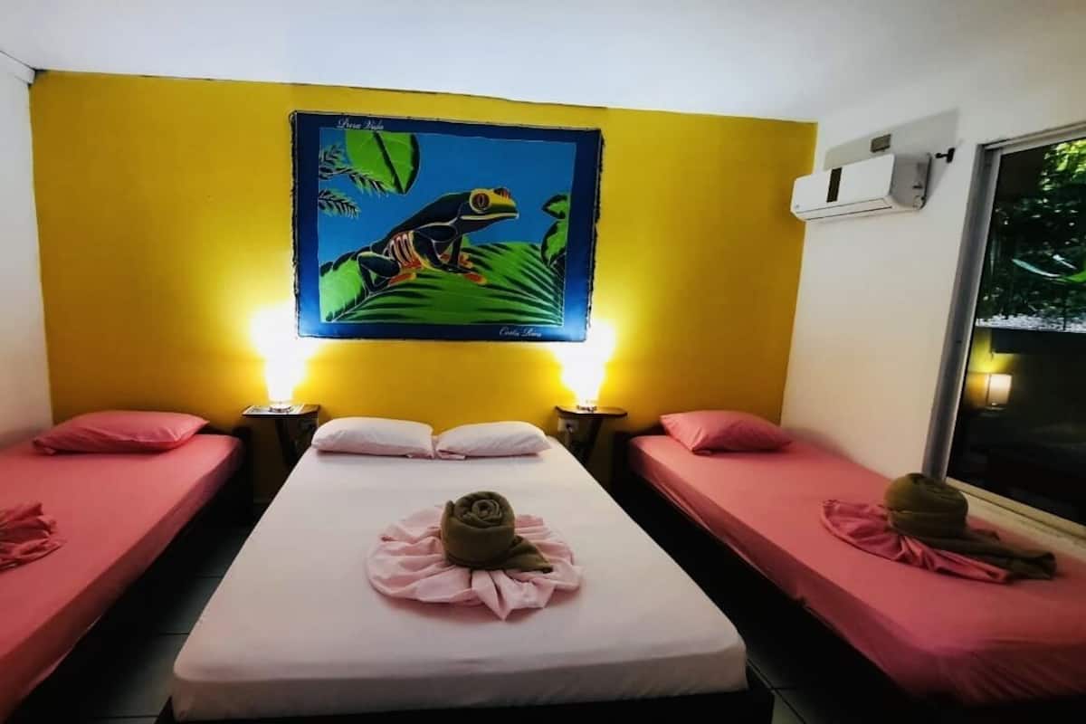 Quarto com duas camas, cada uma com roupa de cama rosa e uma toalha em forma de flor. Há uma pintura de um sapo na parede acima das camas.