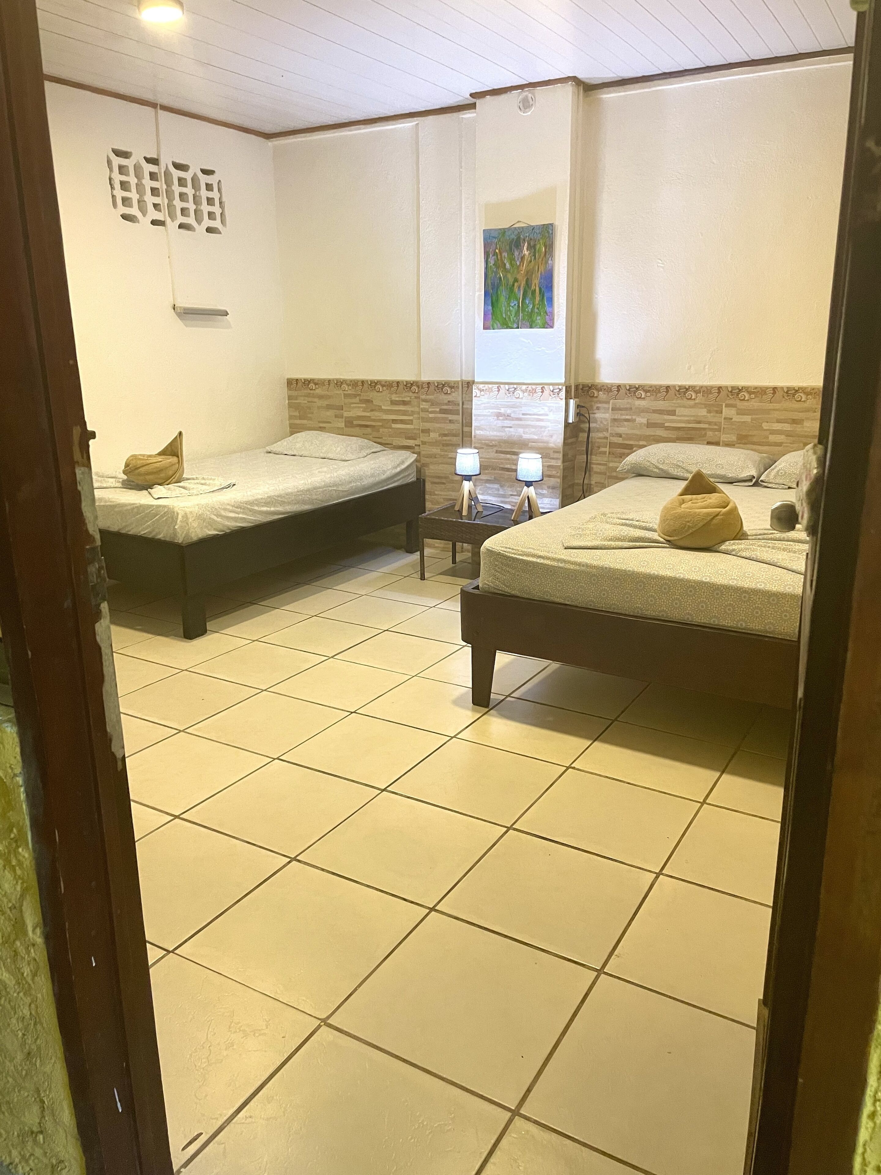 A hotel room with two single beds, a small table with lamps, a painting on the wall, and a tiled floor.