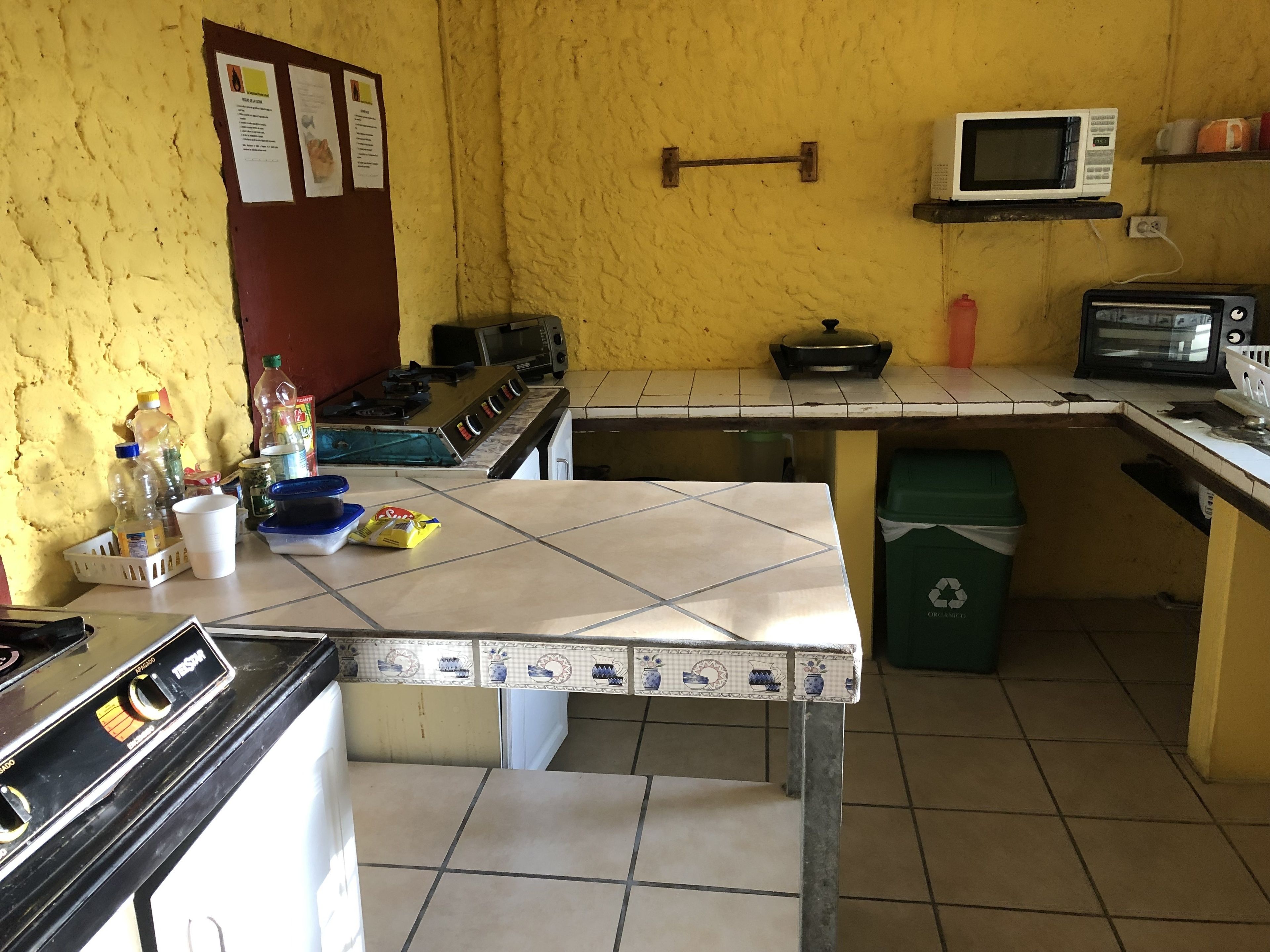 Una cocina con estufa, microondas y lavamanos. Hay varios objetos en la encimera, incluyendo un cubo de basura y una cesta.