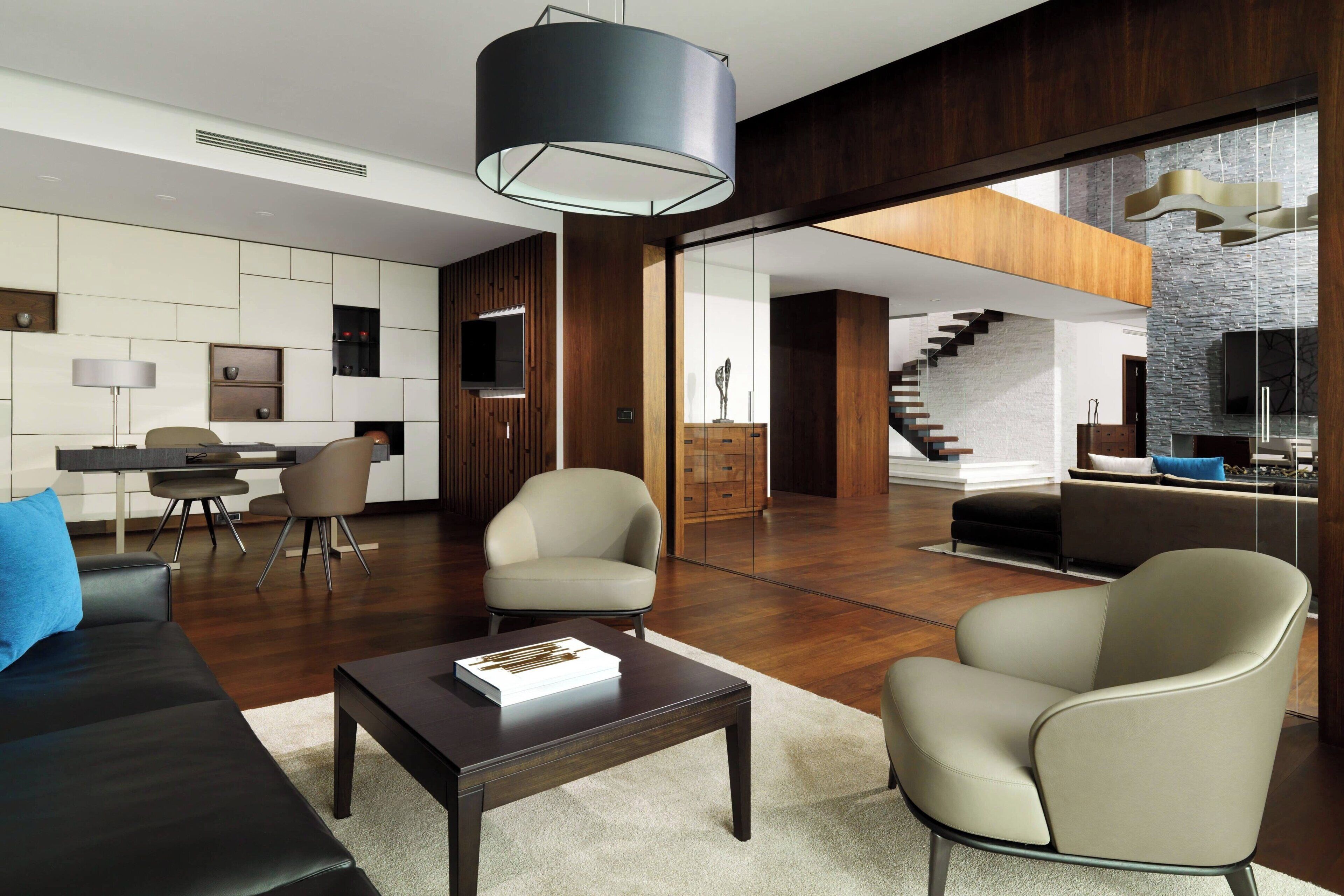 A modern living room with a dark wood floor, a black coffee table, and a beige armchair. There is a staircase leading to an upper level with a stone wall and a wooden ceiling.