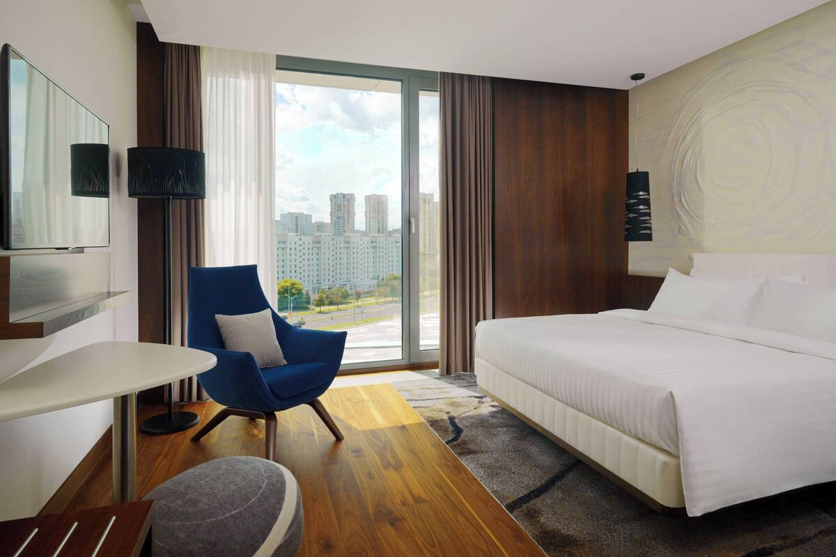 Une chambre d’hôtel avec un lit, une chaise bleue, une télévision et une grande fenêtre donnant sur la ville.