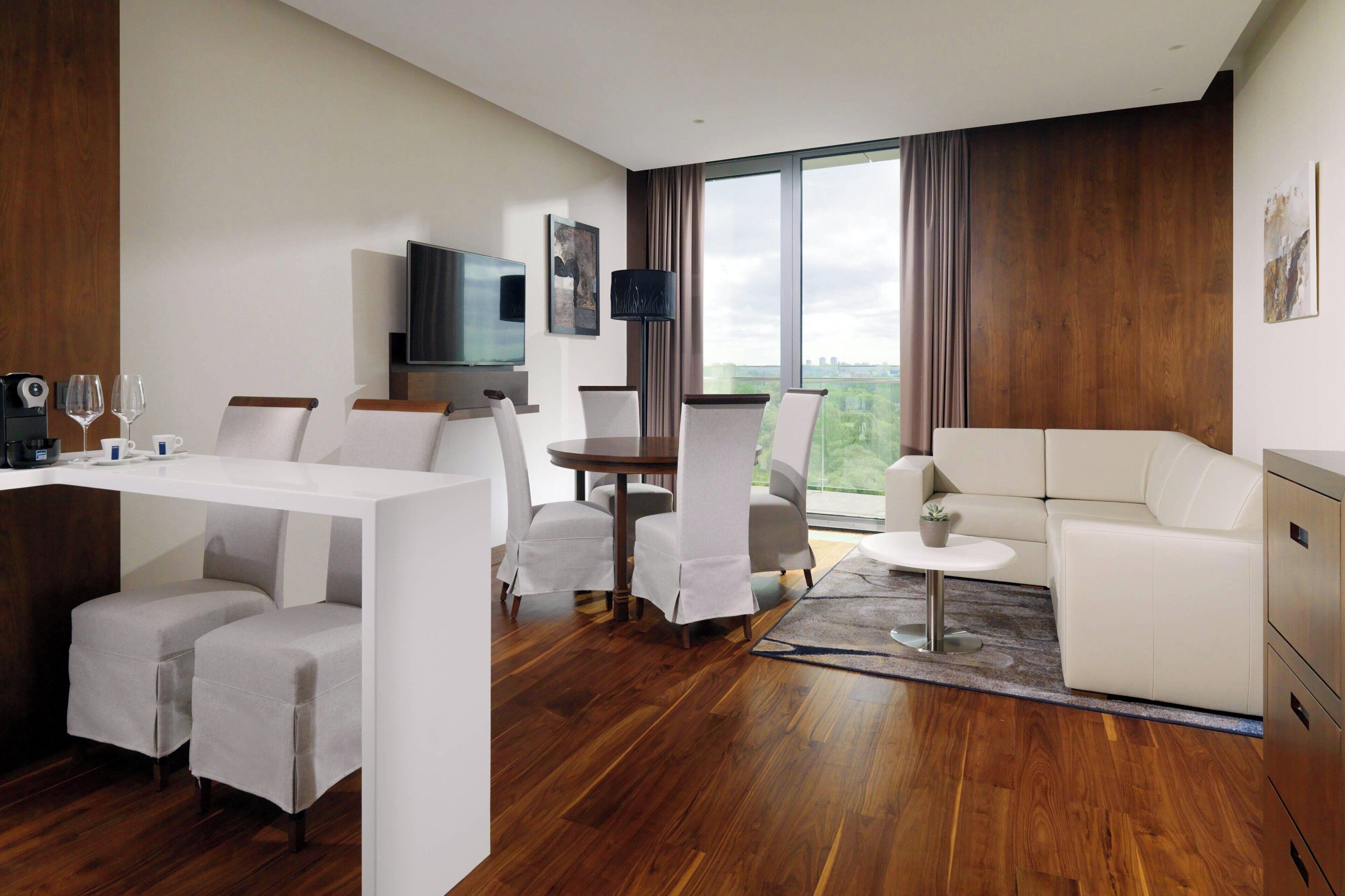 A modern hotel room with a dining area, a sofa, a TV, and a large window offering a city view.