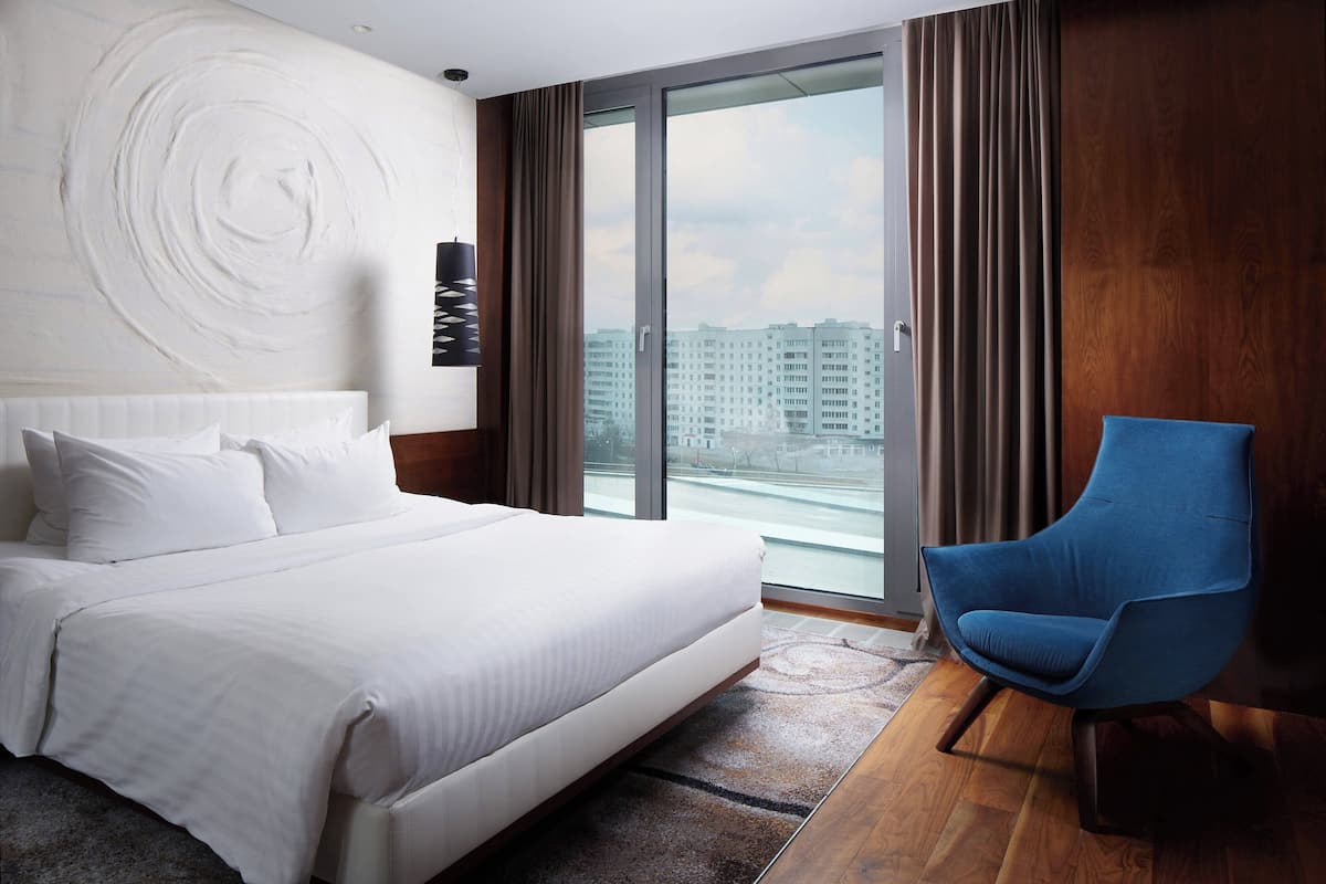 Une chambre d’hôtel moderne avec un grand lit, un fauteuil bleu et une vue sur la ville par une grande fenêtre.