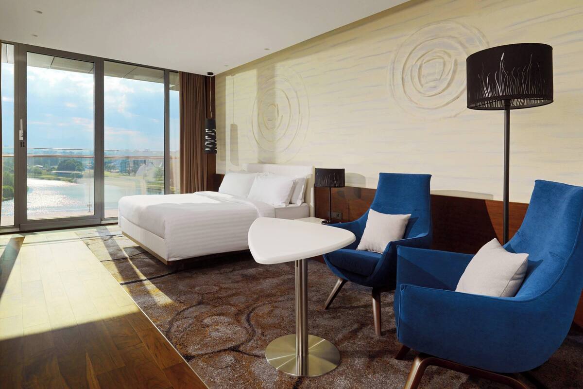 Une chambre d’hôtel avec un lit, deux fauteuils bleus, une petite table et une grande fenêtre donnant sur un plan d’eau.