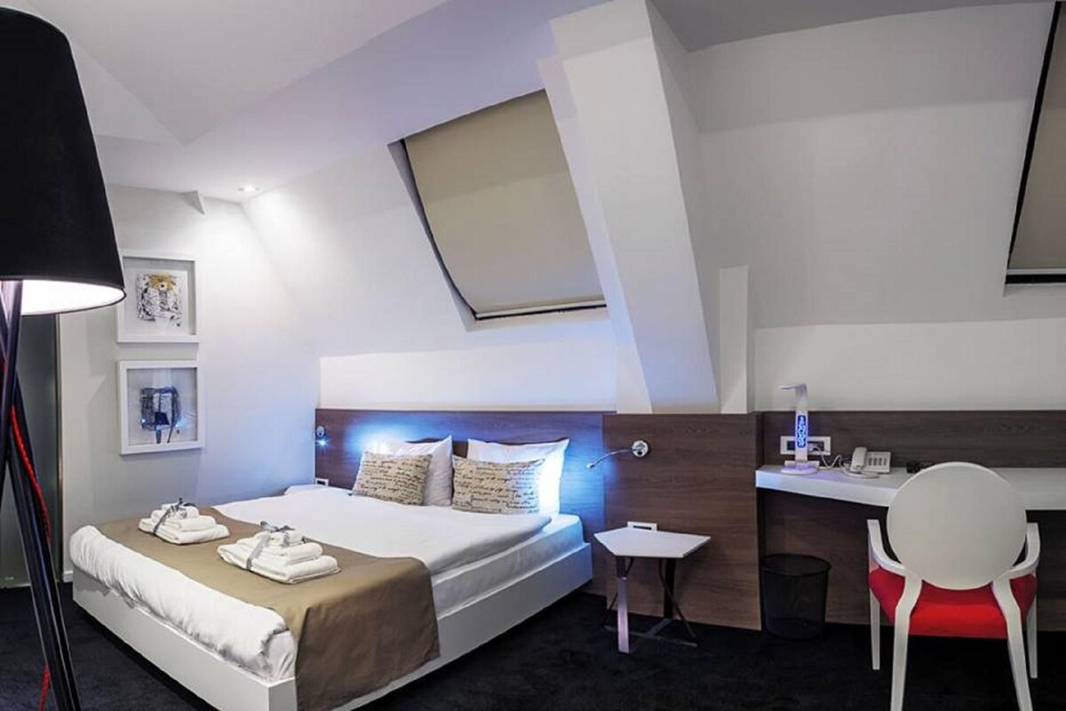 A modern hotel room with a bed, bedside table, desk, chair, and artwork on the wall.