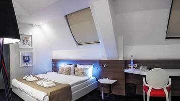 A modern hotel room with a bed, bedside table, desk, chair, and artwork on the wall.