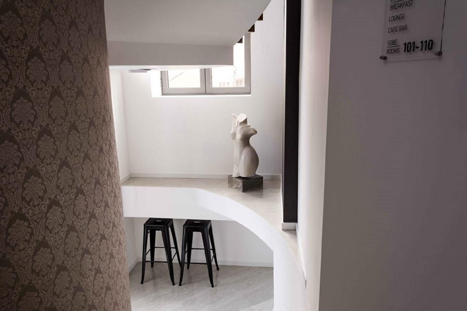 A modern interior with a white wall, a window, and a sculpture. There are two black stools under a white counter.