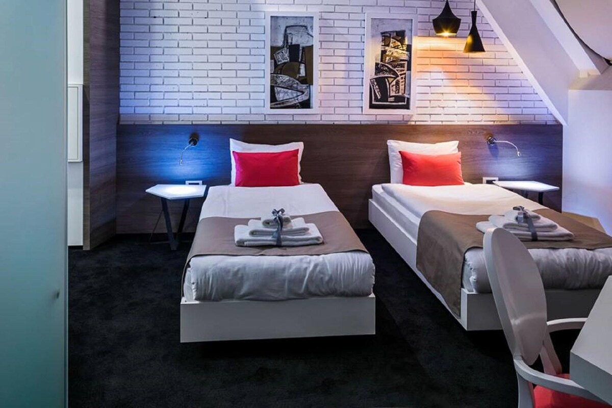 A hotel room with two beds, each with a pillow and a towel. There are wall-mounted artworks and a staircase visible in the background.